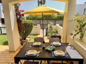 Une terrasse extérieure avec une table dressée pour quatre personnes, entourée de verdure et ombragée par un parasol jaune.