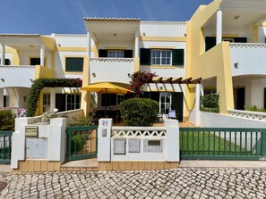A multi-story hotel with a yellow facade, green shutters, and a covered outdoor seating area with an umbrella.
