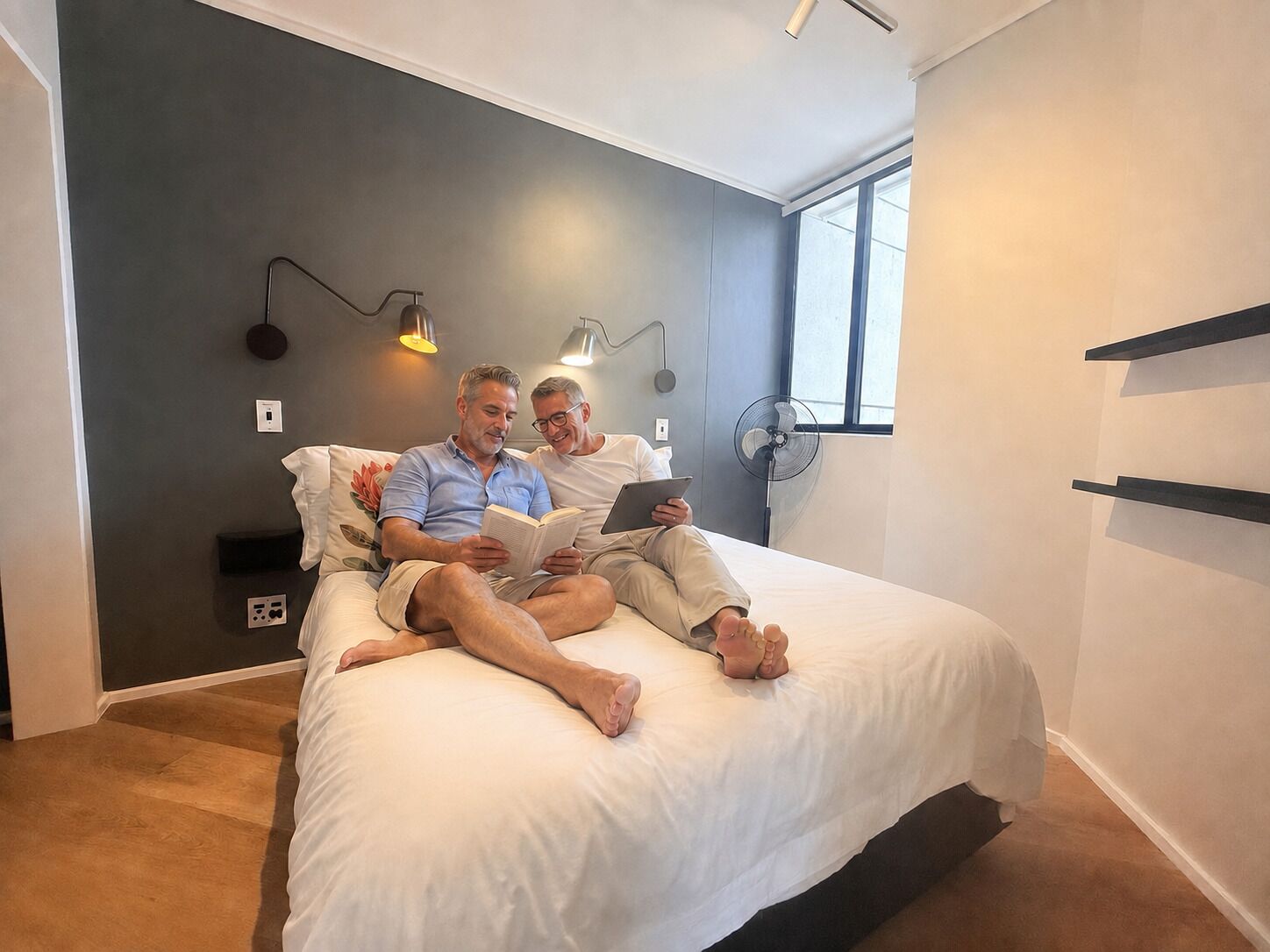 A modern bedroom with a bed, two people reading, a wall-mounted fan, and a window.