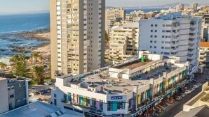 A coastal urban area with a prominent high-rise building and a commercial street with shops. - Urban Elephant Express The Flamingo (Cape Town)