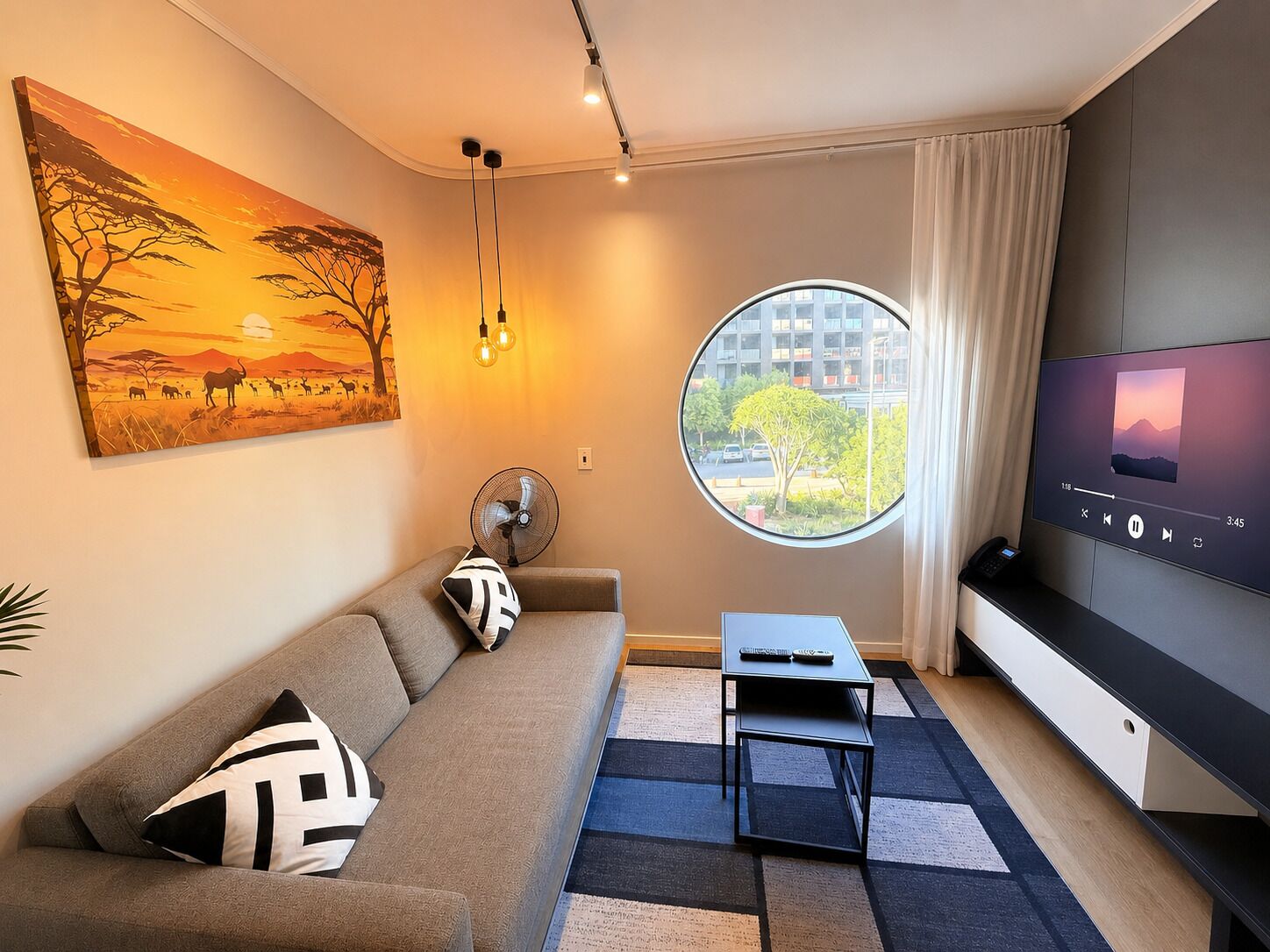 Un salon moderne avec un canapé, une fenêtre ronde, un téléviseur à écran plat et un tableau représentant une scène de savane.
