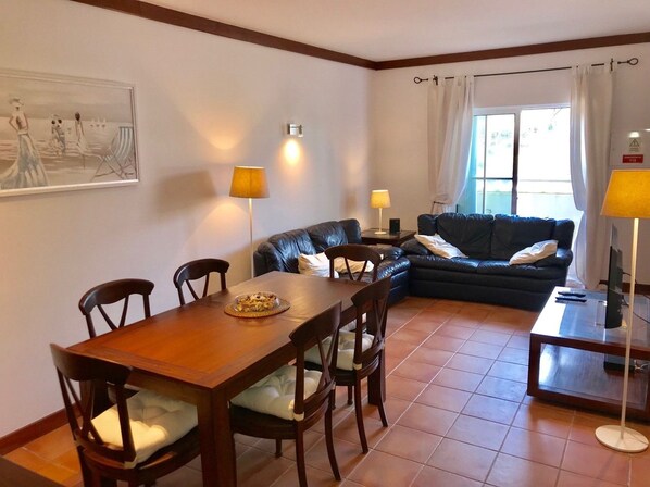 A dining area with a wooden table and chairs, a black leather sofa, and a television on a stand. - 45 Terraços de Benagil (Casa Céu Azul) (Benagil)