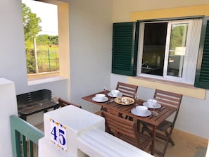 A dining area with a table set for four, a grill, and a window with green shutters. - 45 Terraços de Benagil (Casa Céu Azul) (Benagil)