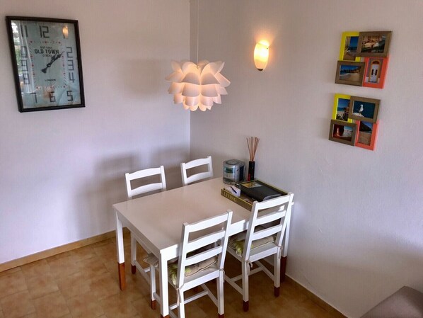 Un coin repas avec une table et des chaises blanches, une horloge murale, un luminaire fixé au mur et une petite étagère sur laquelle se trouvent un livre et une bougie.