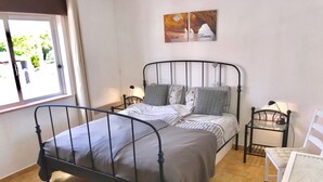 A bedroom with a metal bed frame, a bedside table with a lamp, a chair, and a window with curtains.