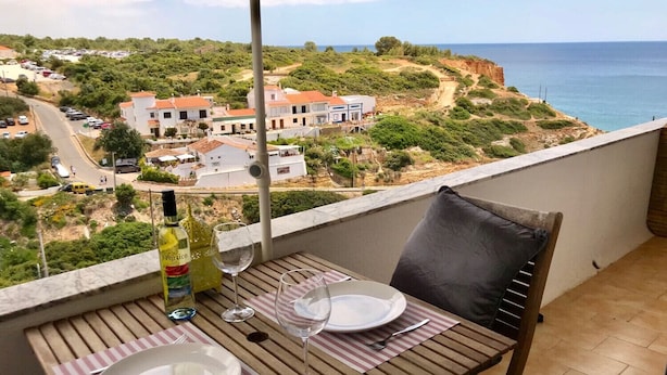 A balcony with a table set for two, overlooking a coastal landscape with buildings and a road.