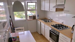 A compact kitchen with a gas stove, sink, and white cabinets. A window offers a view of a parking lot and greenery.