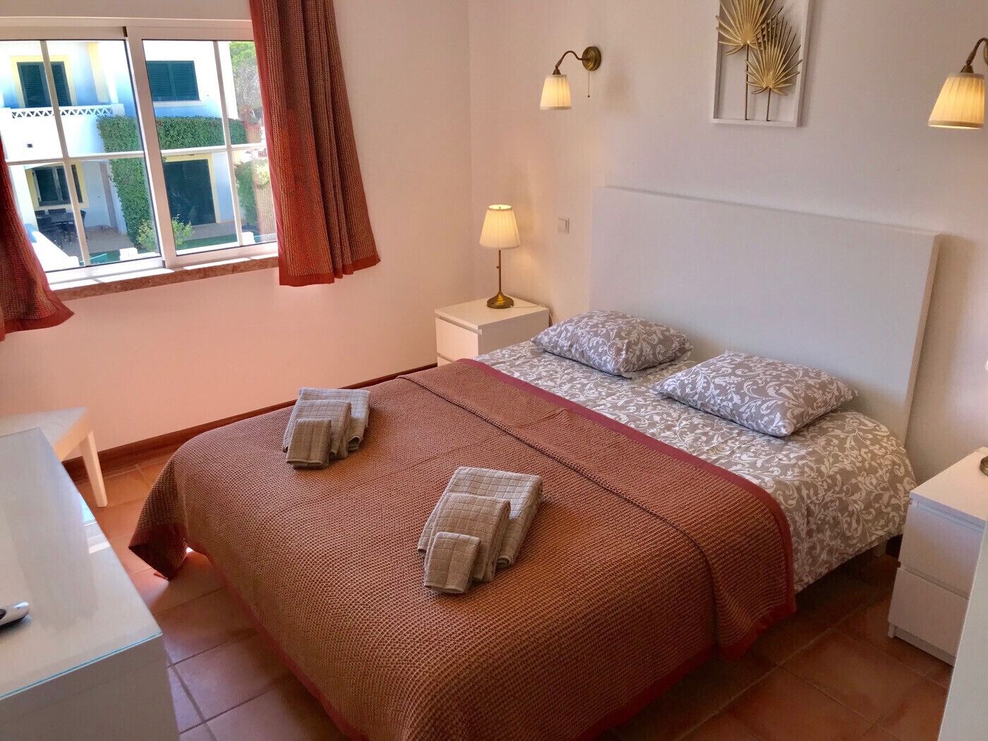 A bedroom with a bed, two bedside tables, a window with curtains, a wall-mounted lamp, and a decorative wall piece.