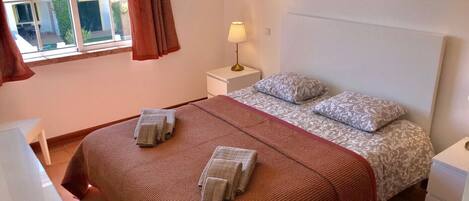A bedroom with a bed, two bedside tables, a window with curtains, a wall-mounted lamp, and a decorative wall piece.