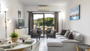 A modern living room with a grey sofa, dining table set with plates and glasses, and a view of a balcony with outdoor furniture. - Casa Colina Sol 611 - Seaview apartment (Carvoeiro)