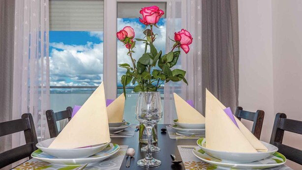 A dining table set with white plates, a vase with pink roses, and a view of the sea through a window. - Apartman Relax (Crikvenica)