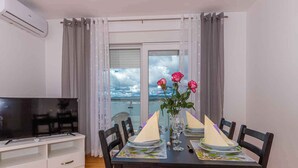 A dining area with a table set for two, a TV on a stand, and a balcony with a view of the sky and clouds. - Apartman Relax (Crikvenica)