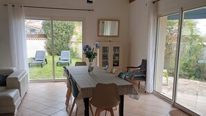 A dining area with a table and chairs, a vase with flowers, and a view of the garden through glass doors. - Quiet family home 10 minutes walk from the sea (Dolus-d'Oléron)