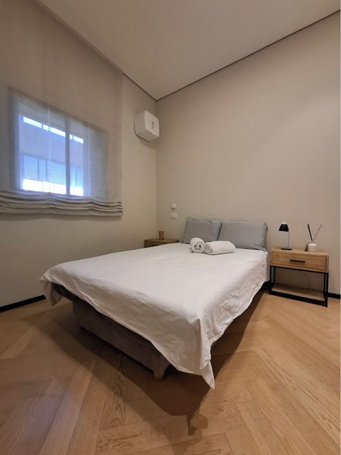 Quarto com cama, mesa de cabeceira, luminária e janela com cortinas.