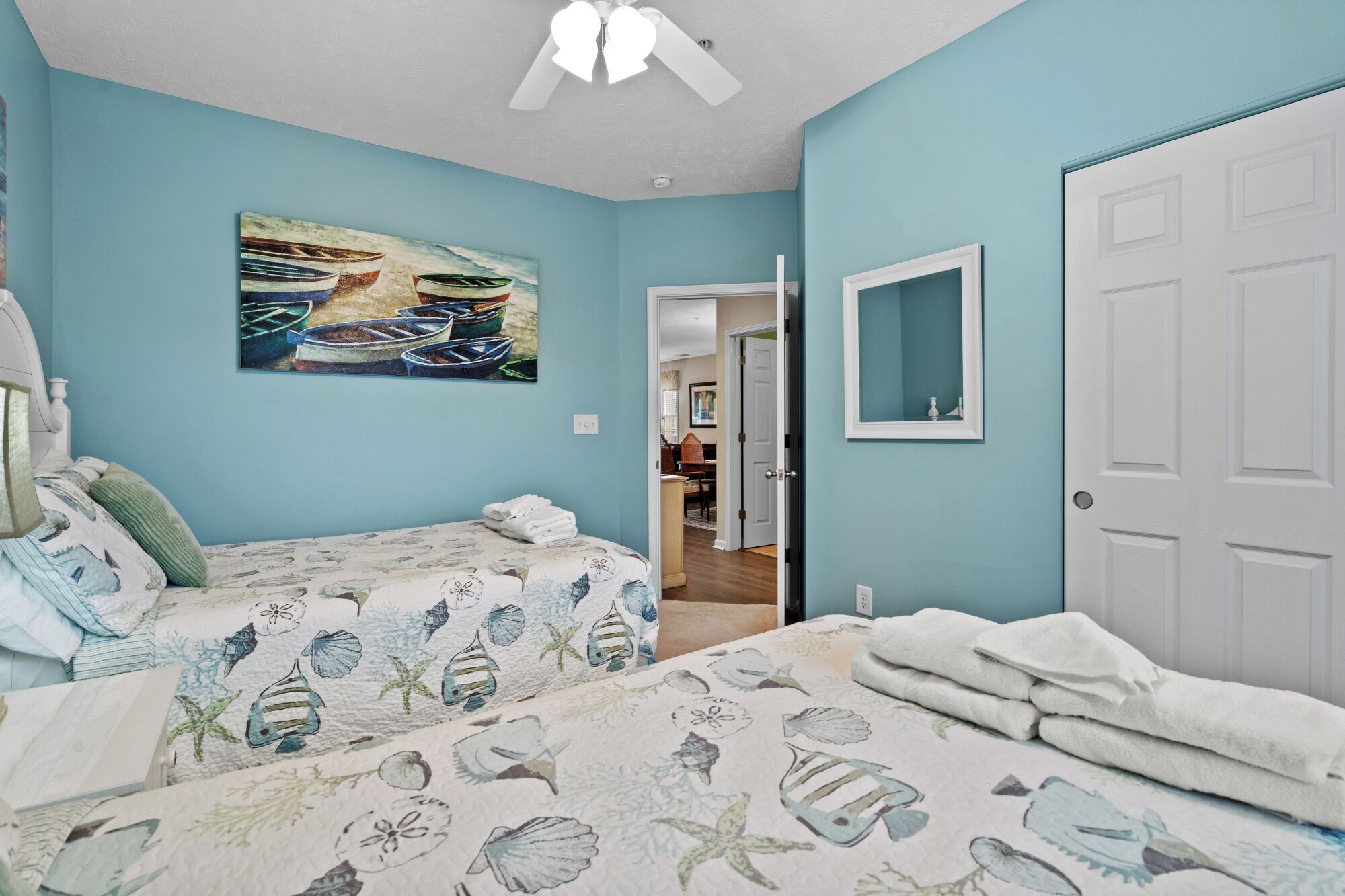 Een slaapkamer met twee bedden, een plafondventilator, een schilderij van boten en een deur die uitkomt in een gang.