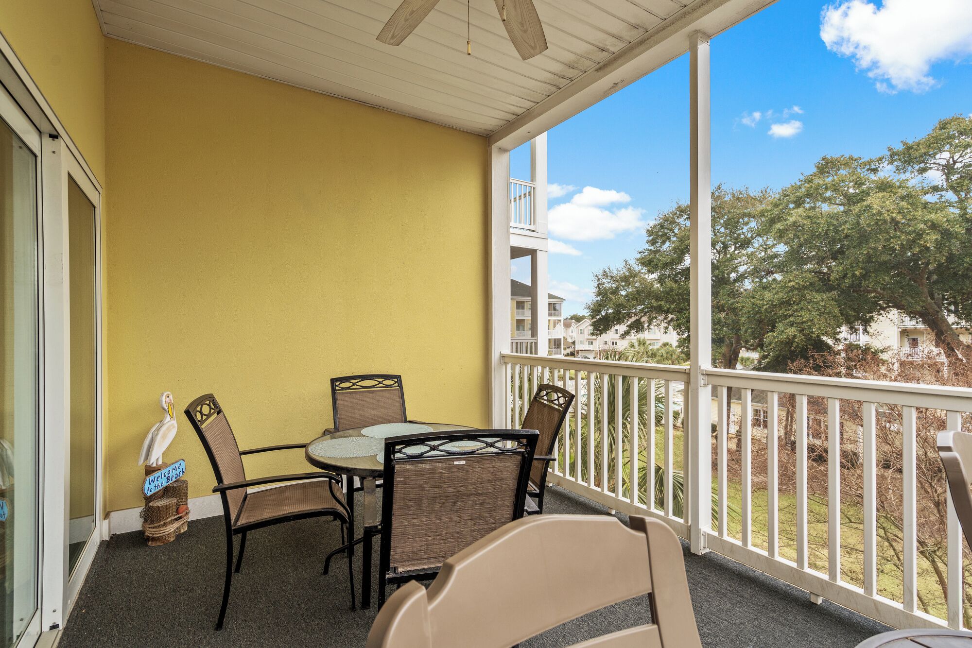 A balcony with a yellow wall, a table with chairs, and a view of a residential area with trees and buildings.