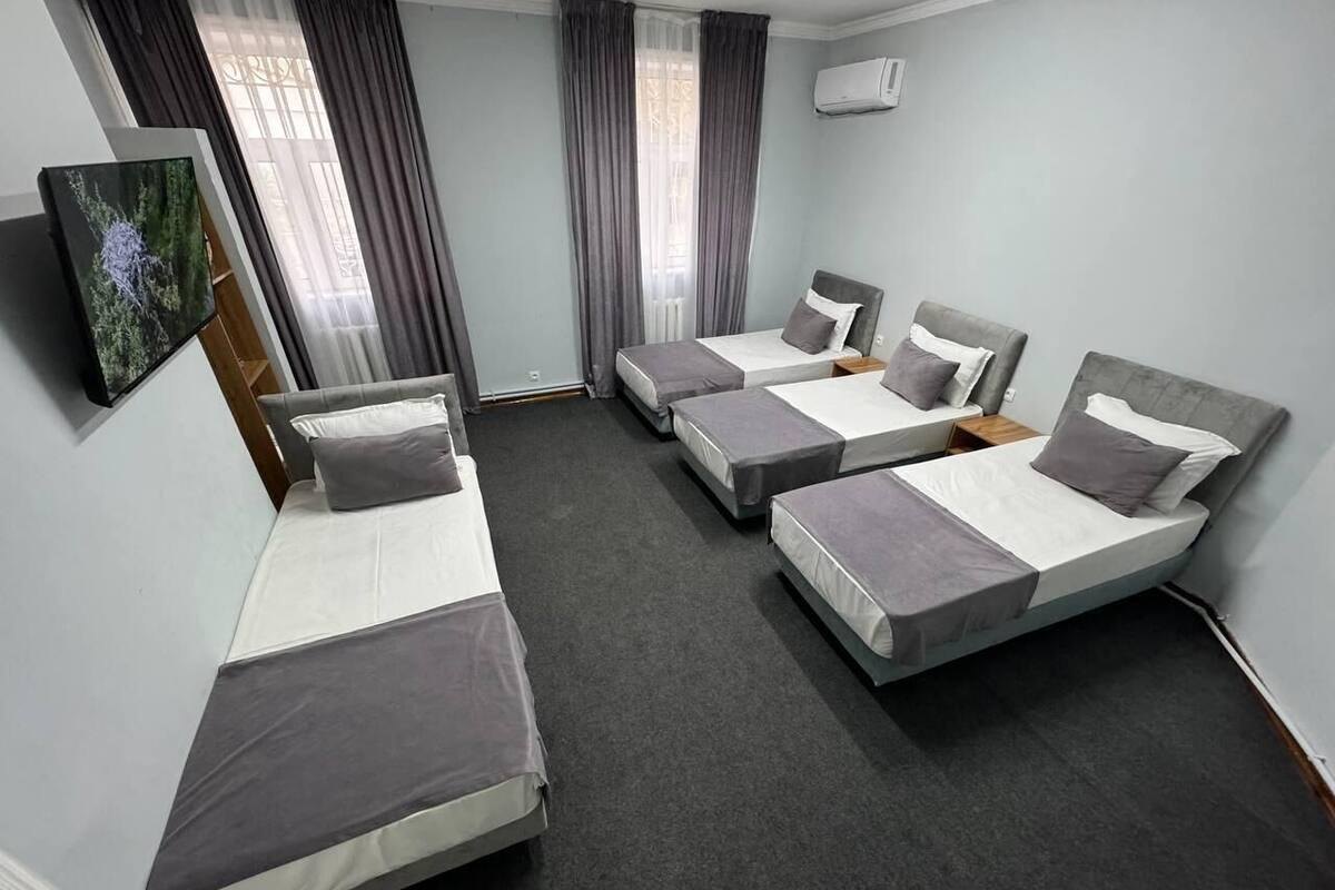 A hotel room with four single beds, grey bedding, a grey carpet, a painting on the wall, and a window with curtains.