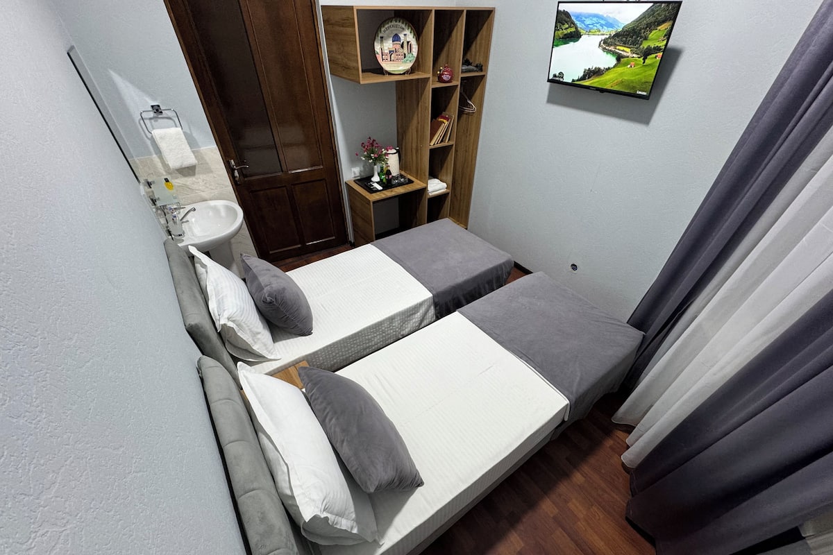 A small, compact bedroom with two single beds, a wooden wardrobe, a small table with a mirror, and a television on the wall.