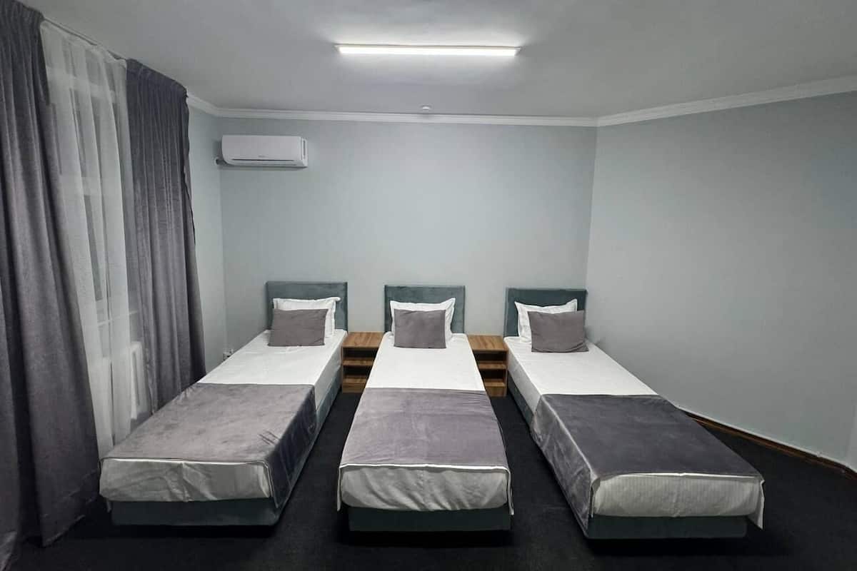 A hotel room with three single beds, a ceiling light, an air conditioning unit, and a window with curtains.