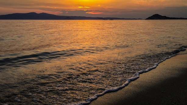 Uma praia com um pôr do sol sobre a água, ondas suaves beijando a costa e montanhas ao longe.