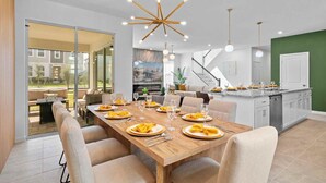 A modern dining area with a wooden table set for six, a kitchen with white cabinets, and a view of a residential building through large glass doors. - 2315  Amazing house 6/5 W/Pool Solara Resort (Kissimmee)