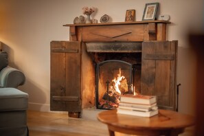 A fireplace with a mantel, a fire burning inside, and a stack of books on a table in front.