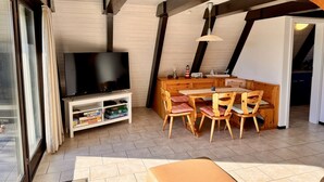 A cozy room with a TV on a stand, wooden furniture, and a window with curtains. - Nurdachhaus in Burhave (Butjadingen)