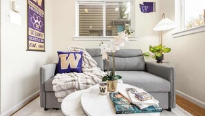 A cozy living room with a grey sofa, a round coffee table with books and candles, a potted orchid, and a 'W' pillow.