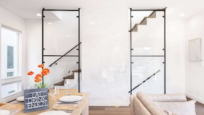 Een moderne eetruimte met een houten tafel voor twee personen, een glazen scheidingswand en een trap met een metalen frame.