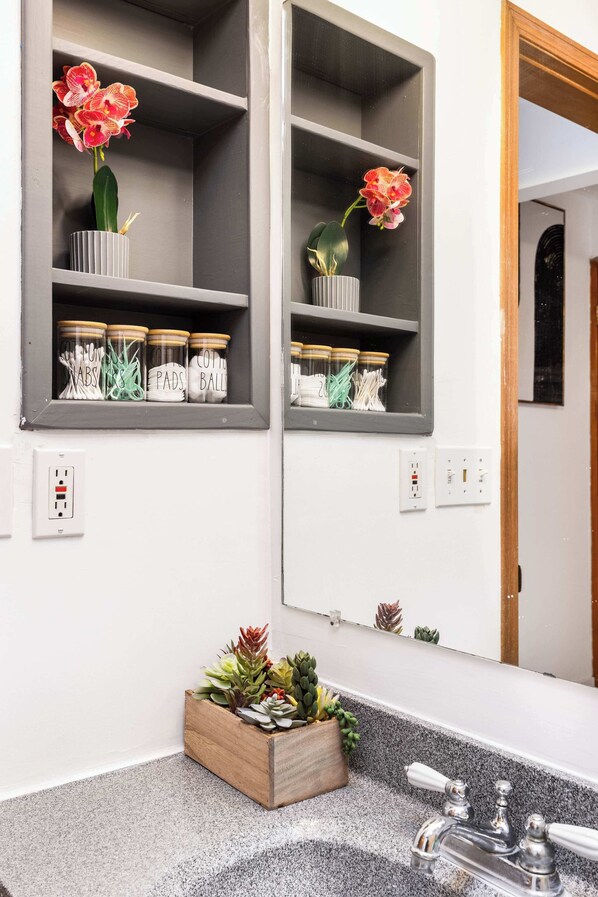 A bathroom with a granite countertop, a wooden planter with succulents, a mirror, and a shelf with jars and flowers.
