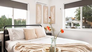 A bedroom with a bed, a nightstand, a vase with flowers, and a window with a view of houses. - Modern 2BR • Near DT + Stadiums • Parking (Seattle)