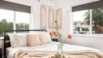 Ein Schlafzimmer mit Bett, Nachttisch, Blumen in einer Vase und einem Fenster mit Blick auf Häuser.