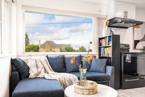 A living room with a sofa, a coffee table, a bookshelf, and a kitchen area with an oven.