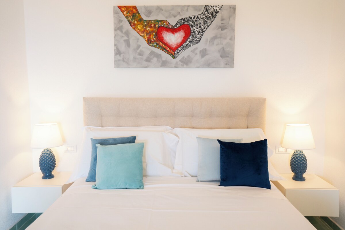 A neatly made bed with white and blue pillows, a beige headboard, and a wall art piece above it.