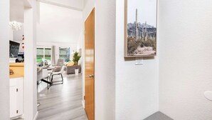 A hallway leading to a living area with a wooden door, a framed picture on the wall, and a view of the outdoors.