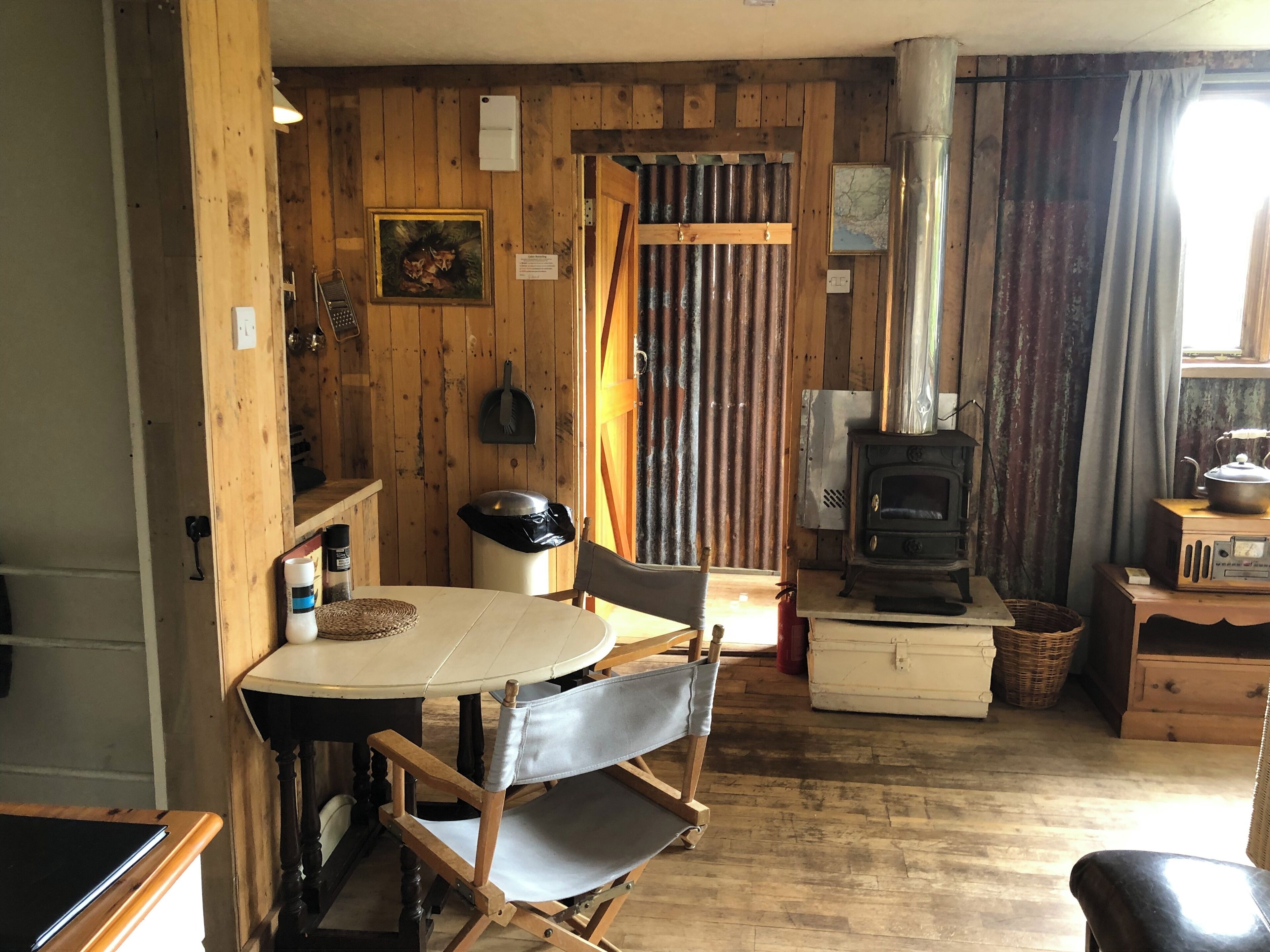 A rustic wooden interior with a dining table, chairs, a stove, and a trash bin.