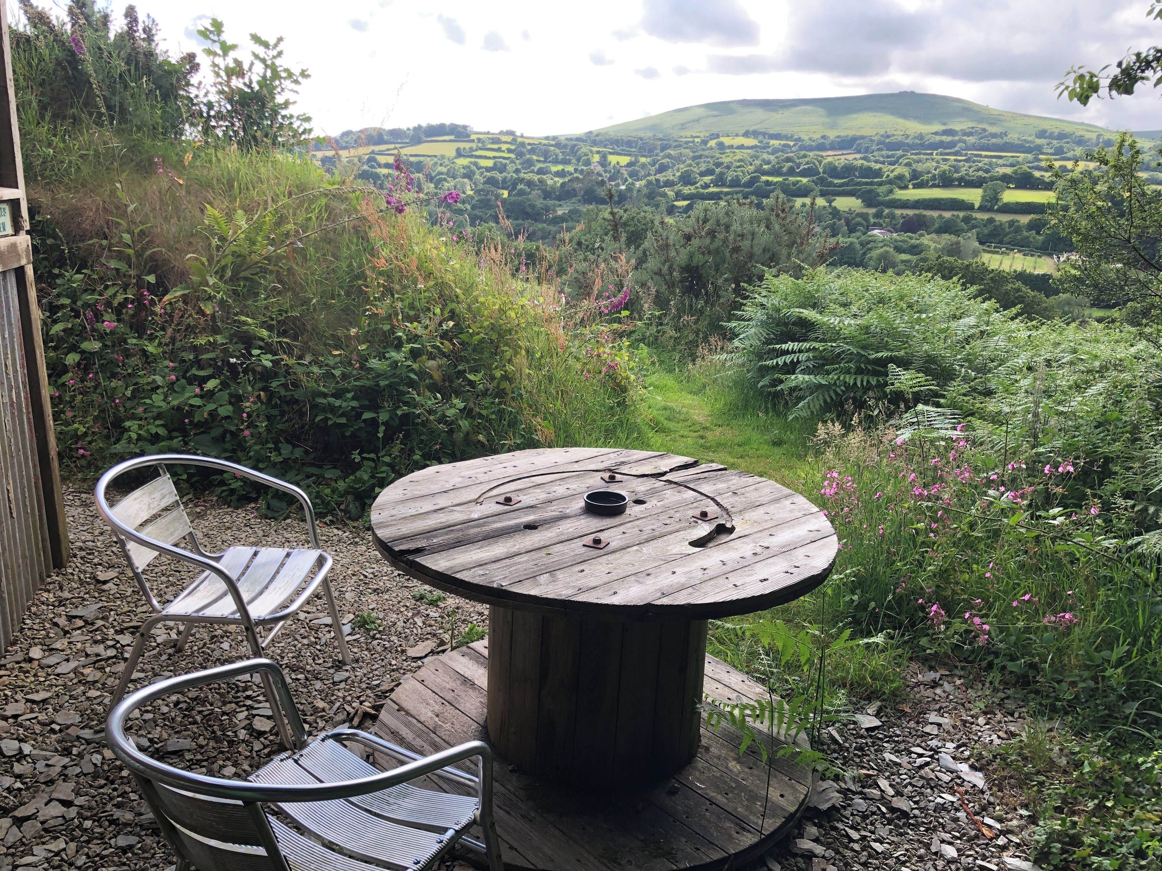 A wooden table with metal chairs in a garden with a view of rolling hills.