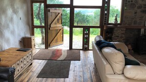 A rustic cabin interior with a wooden floor, a white sofa, a wooden cabinet, and a large window with a view of greenery. - Monument Field South Brent (South Brent)