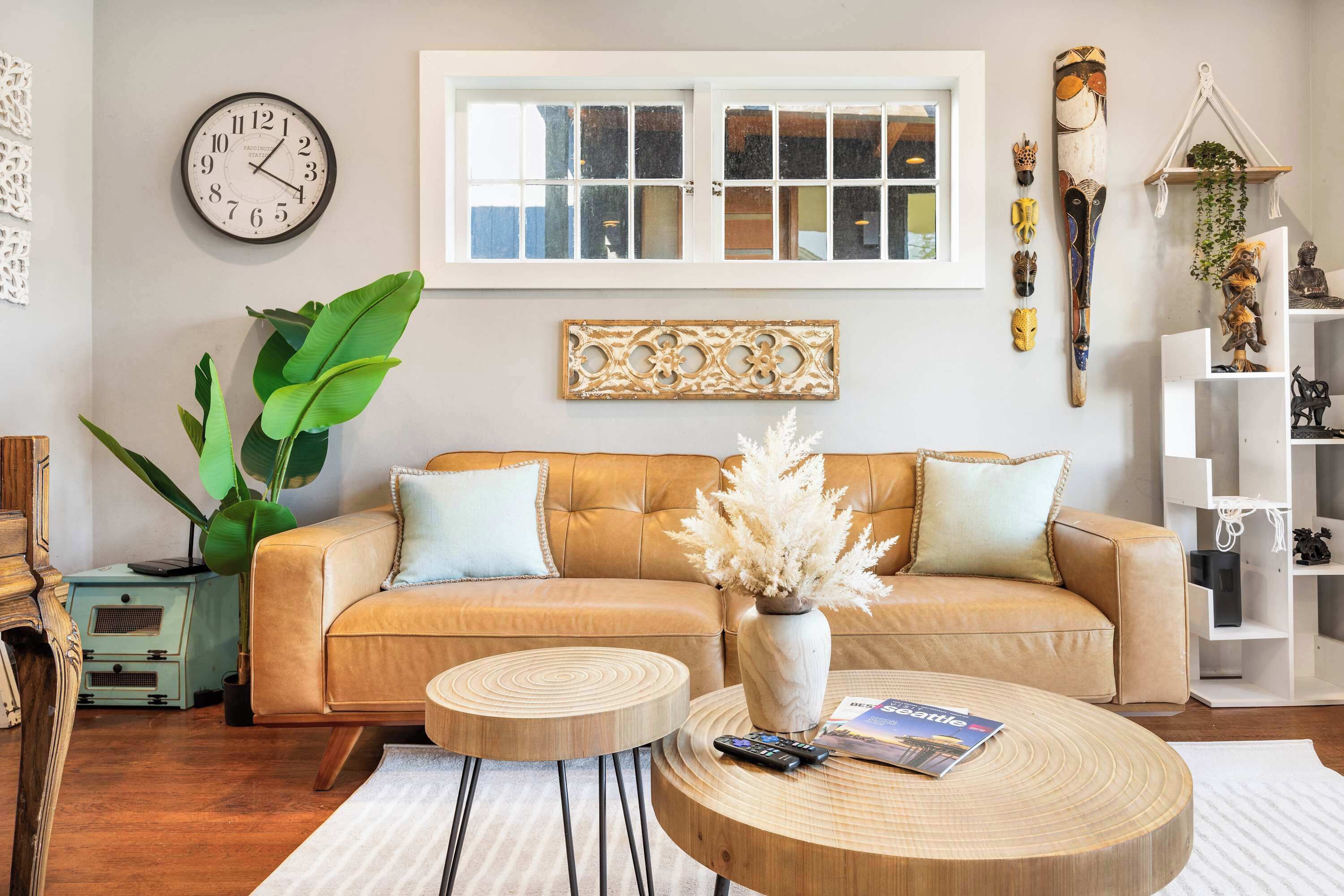 Ein Wohnzimmer mit beiger Couch, zwei runden Holztischen, einem großen Fenster, einer Wanduhr und Dekorationselementen.