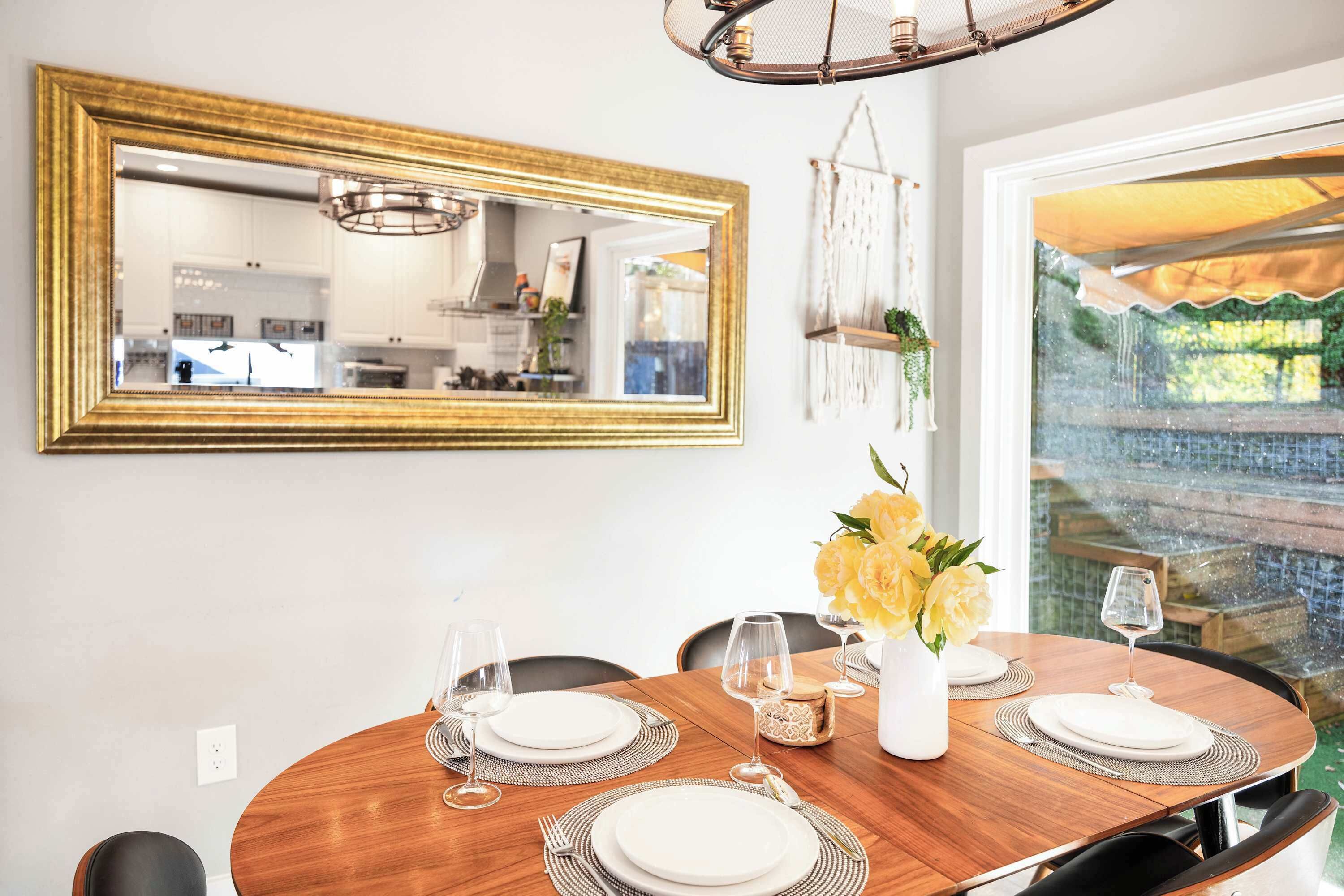 A dining area with a wooden table set for four, a gold-framed mirror, and a view of a garden through a glass door.