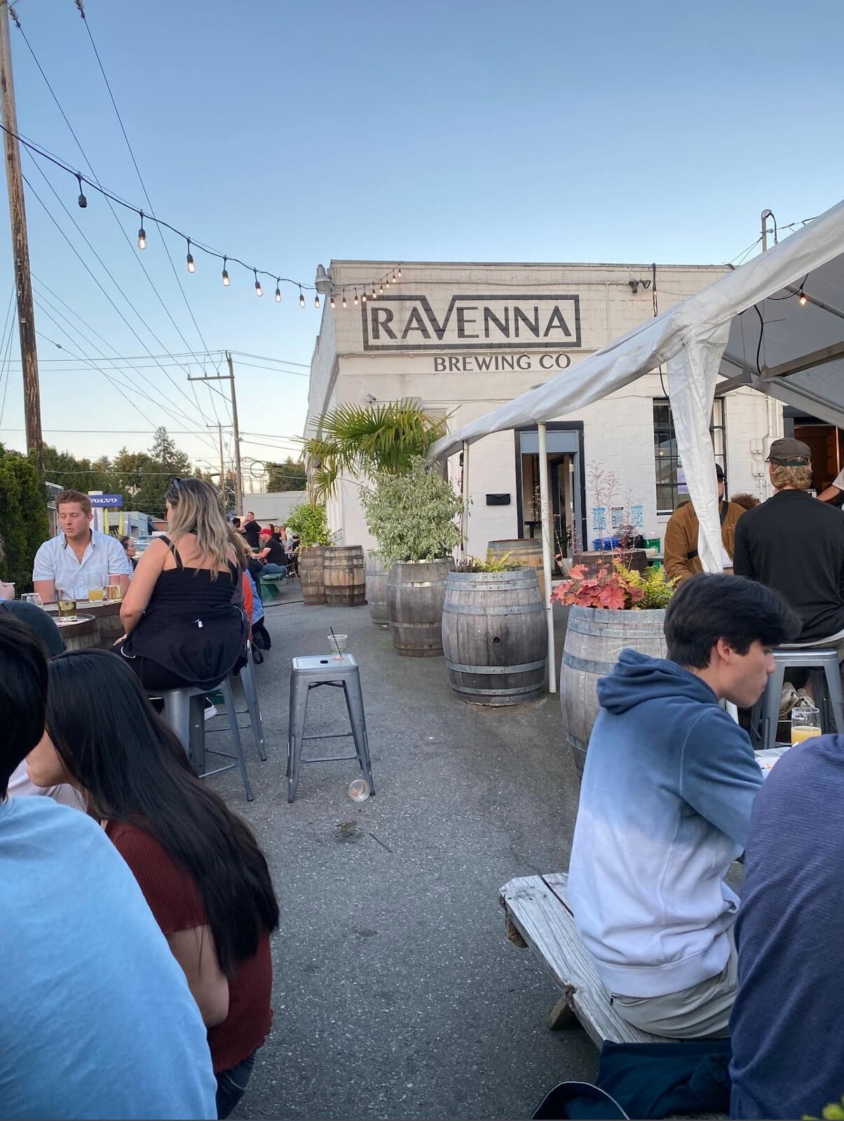 An outdoor seating area at Ravenna Brewing Co. with patrons seated at tables, surrounded by wooden barrels and string lights.