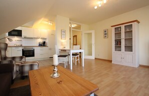 A compact kitchen with white cabinets, a dining area with a wooden table, and a built-in cabinet with glass doors. - Ferienwohnung in Burhave (Butjadingen)