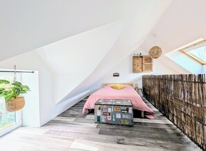 A bedroom with a sloped ceiling, a bed with a pink bedspread, a hanging plant, a wooden cabinet, and a large window.