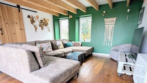 A living room with a sectional sofa, a small table, and a TV stand. There are wooden beams on the ceiling and a green wall.