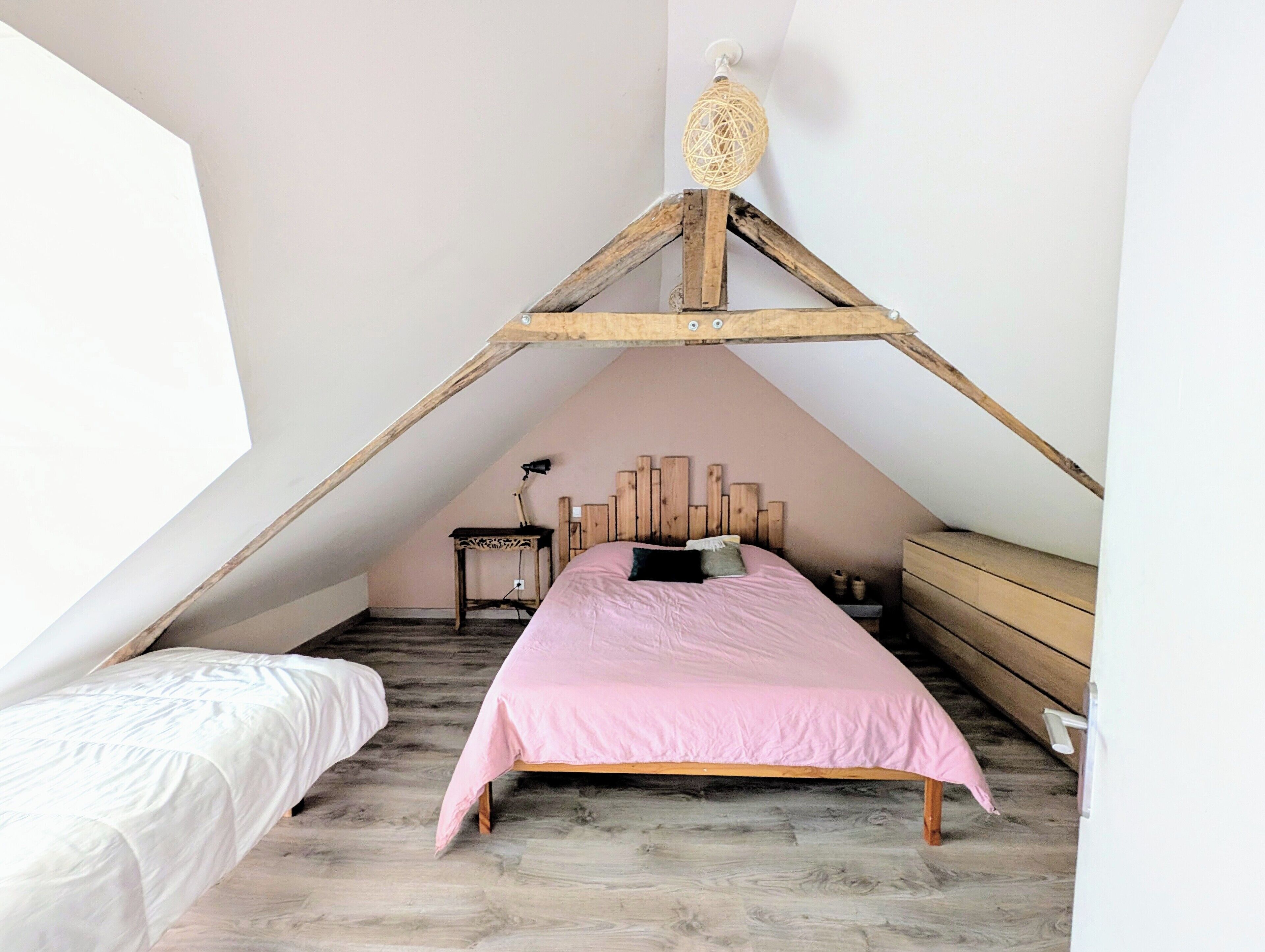 A bedroom with a sloped ceiling, a bed with a pink bedspread, a wooden headboard, a small table, and a hanging light fixture.
