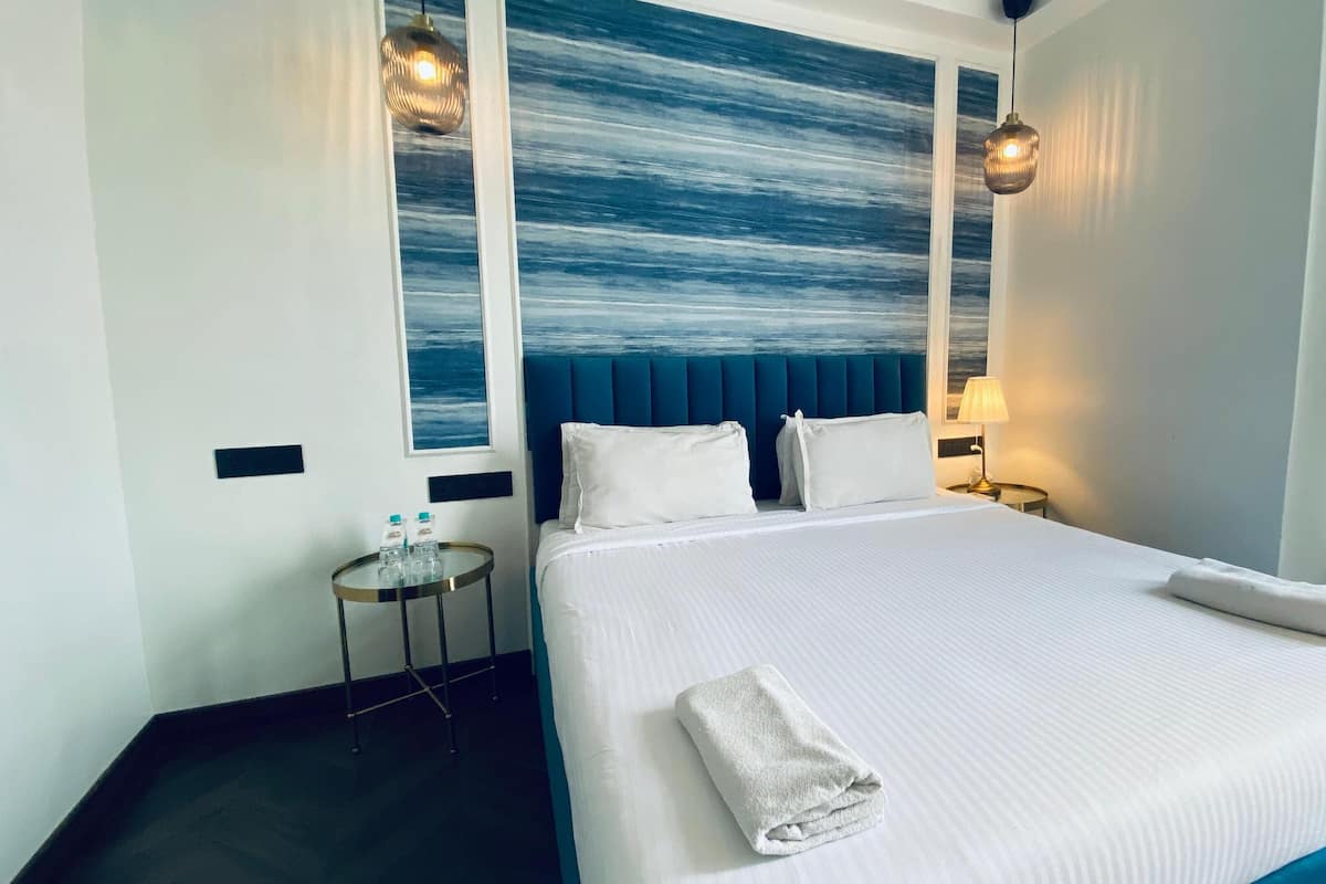 Habitación de hotel con una cama grande, cabecera con panel azul, dos lámparas de noche y una mesita redonda con una botella de agua.