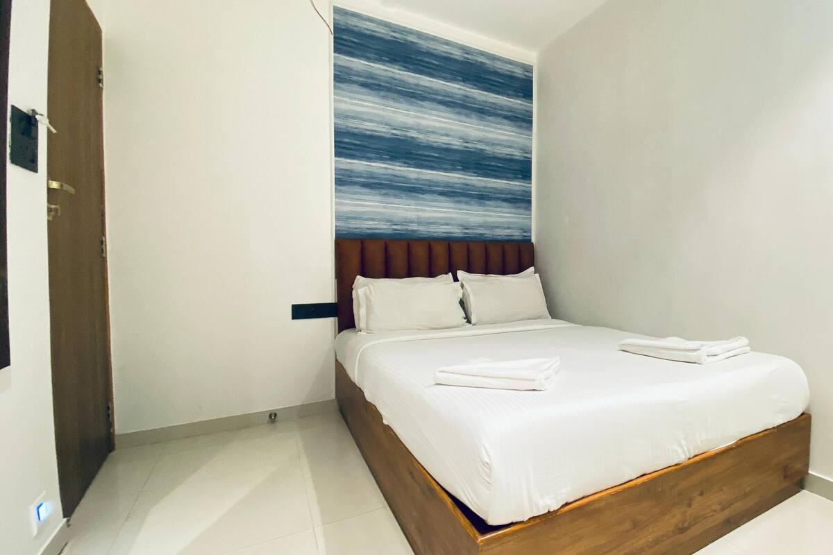 Una cama bien hecha en una habitación de hotel, con cabecera de madera, ropa de cama blanca y un cuadro abstracto azul en la pared.