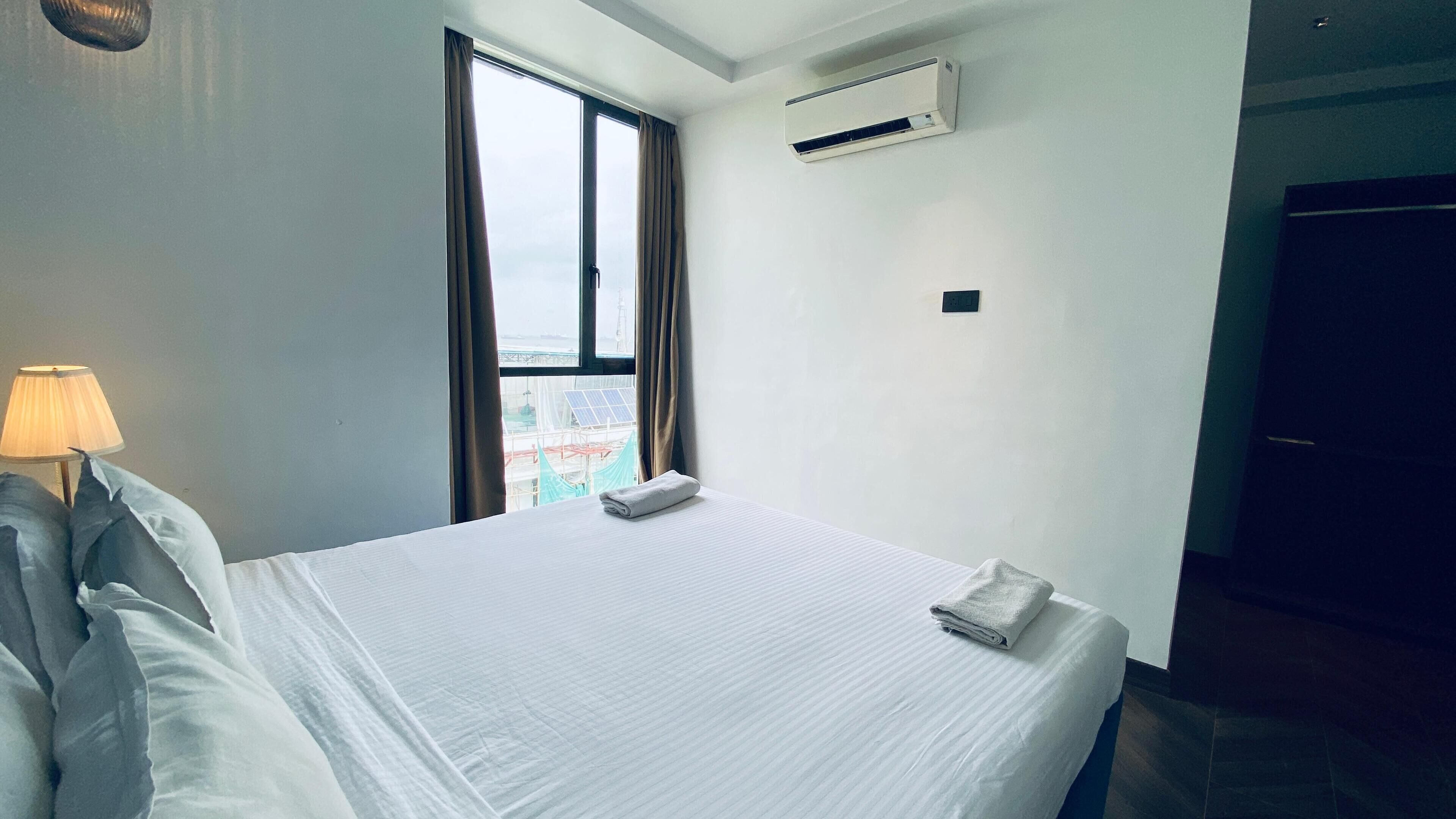 A hotel room with a bed, a window with curtains, an air conditioning unit, and a lamp.