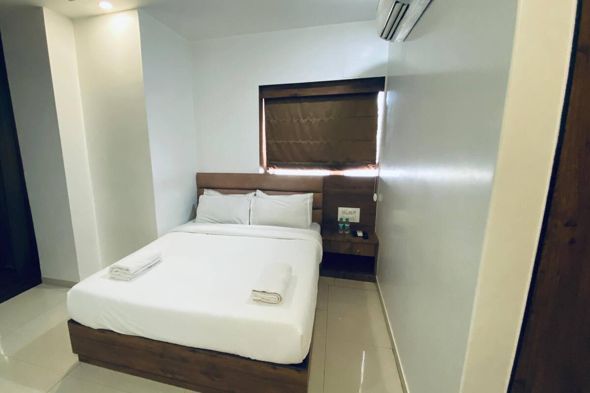 Una cama bien hecha con sábanas y almohadas blancas, cabecera de madera, mesita de noche con lámpara y un aire acondicionado de pared.