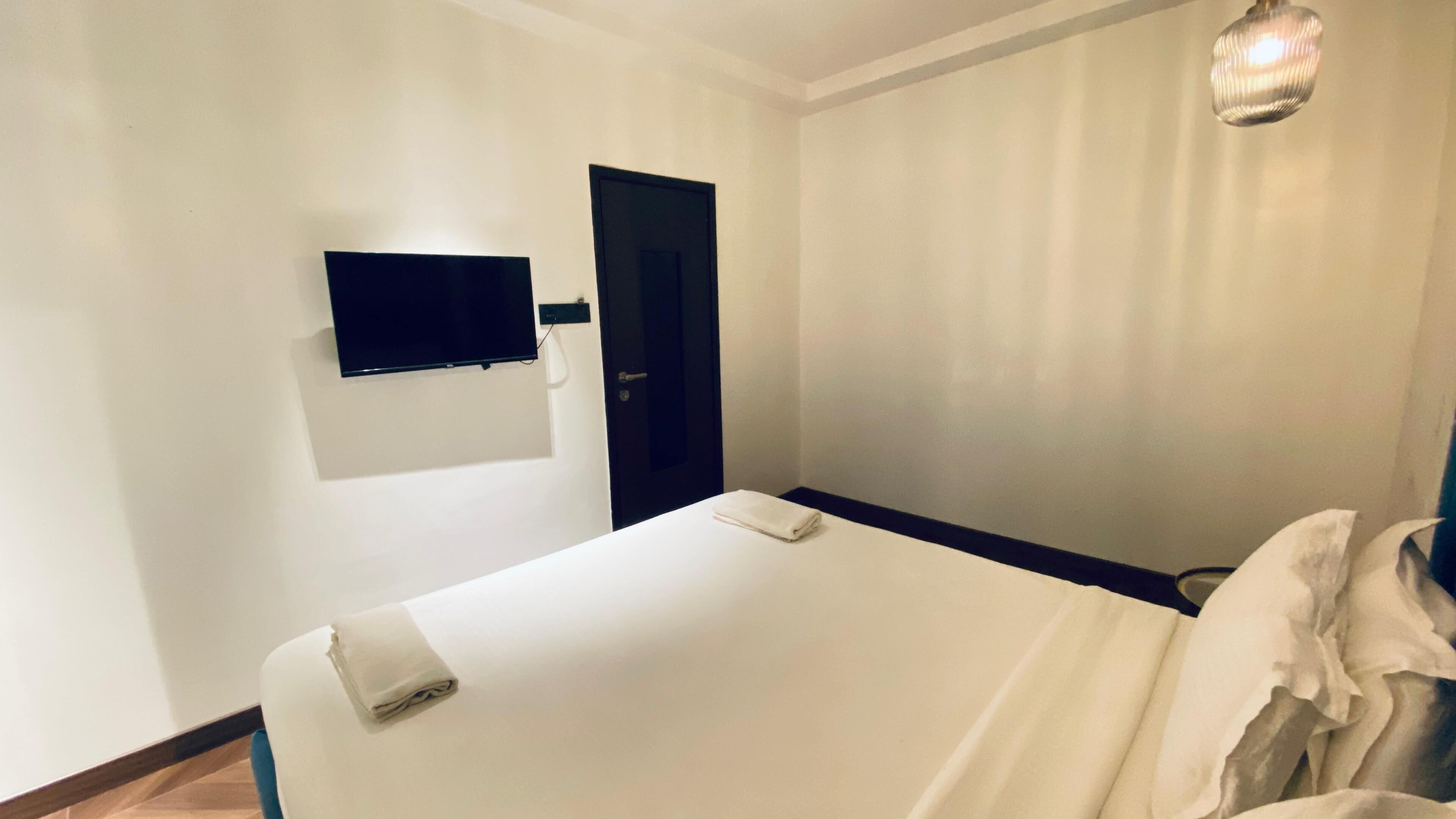 A hotel room with a bed, a flat-screen TV mounted on the wall, a door, and a light fixture.